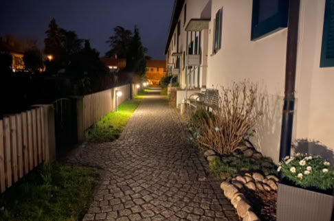 FHG am Hörnli, Gepflasterter Gehweg neben einem Gebäude mit Fenstern, beleuchtet von Laternen, umgeben von Pflanzen und einem Holzzaun, bei Nacht.