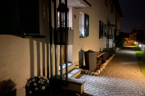 FHG am Hörnli, Beleuchteter Hauseingang bei Nacht mit Pflasterweg, Blumen und Bank.