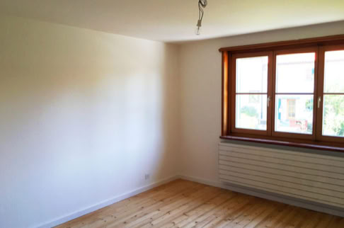 FHG am Hörnli, Leerer Raum mit Holzboden, weiß gestrichenen Wänden, einem großen Fenster mit Holzrahmen und einem Heizkörper darunter.